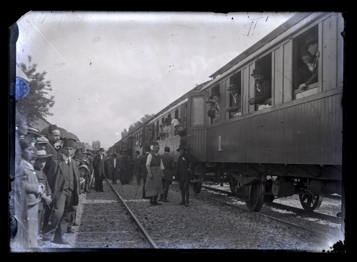 Tren oprit în gară, cu călători la ferestre și oameni pe peron